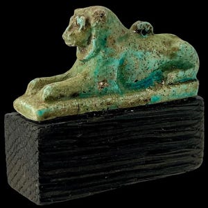 Op de afbeelding: Een turquoise en groene leeuwensculptuur, mogelijk oud, rustend op een donker, getextureerd houten blok. De leeuw ligt in een liggende houding, met gedetailleerde kenmerken. De sculptuur staat tegen een zwarte achtergrond.