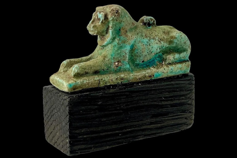 Op de afbeelding: Een turquoise en groene leeuwensculptuur, mogelijk oud, rustend op een donker, getextureerd houten blok. De leeuw ligt in een liggende houding, met gedetailleerde kenmerken. De sculptuur staat tegen een zwarte achtergrond.