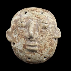 Pode incluir: Uma escultura desgastada, esbranquiçada, de um rosto humano com olhos fechados, um nariz proeminente e uma boca pequena. A escultura tem vários pequenos orifícios e está sobre um fundo preto. A peça parece ser um artefato antigo.