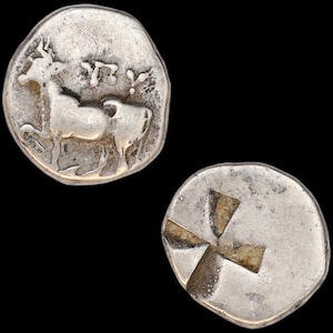 May include: Two silver ancient coins. One coin features a detailed image of an animal, possibly a bull or cow, with text above. The other coin has a cross design. Both coins are on a black background.