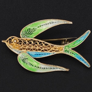 Pode incluir: Broche de pássaro dourado com detalhes intrincados em filigrana. As asas e a cauda apresentam detalhes em esmalte verde e azul. Uma pequena pérola está localizada perto da cabeça do pássaro. Em um fundo preto.