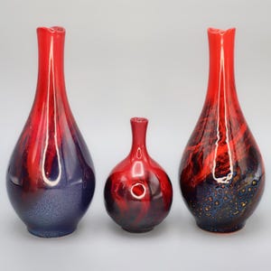 May include: Three decorative ceramic vases in varying sizes. The vases feature a glossy finish with a color gradient of red and dark blue. The smallest vase is round, while the other two are elongated with narrow necks.