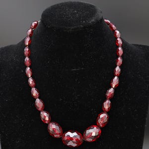 Op de afbeelding: Een gefacetteerde, dieprode kralenketting, gepresenteerd op een zwarte fluwelen buste. De ketting heeft een gegradueerd ontwerp met grotere kralen in het midden en kleinere kralen naar de sluiting toe. De kralen hebben een sprankelend, edelsteenachtig uiterlijk.