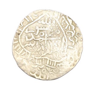 Puede incluir: Una moneda de plata con intrincadas escrituras y dise&ntilde;os &aacute;rabes. La moneda tiene forma circular con un borde ligeramente irregular, y los detalles est&aacute;n en relieve sobre el fondo. Parece ser una moneda antigua.