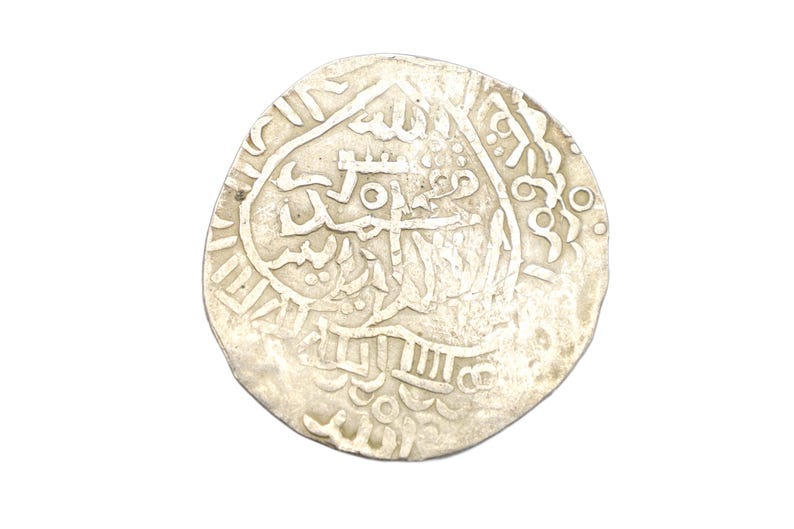 Puede incluir: Una moneda de plata con intrincadas escrituras y dise&ntilde;os &aacute;rabes. La moneda tiene forma circular con un borde ligeramente irregular, y los detalles est&aacute;n en relieve sobre el fondo. Parece ser una moneda antigua.