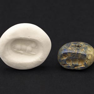 May include: A white, oval-shaped mold with a carved animal design next to a small, blue-grey stone with carved markings. The stone has a hole drilled through it. Both objects are on a black background.
