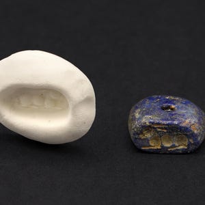 May include: A white, oval-shaped object with a recessed design next to a small, dark blue stone with a hole. The stone has a rough texture and appears to have markings. The objects are set against a black background.