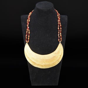Può includere: Una collana a forma di mezzaluna con un grande ciondolo in conchiglia lucida color beige chiaro, sospeso a due fili di piccole perline ovali marroni infilate su un sottile cordino rosso, creando un aspetto stratificato e strutturato.