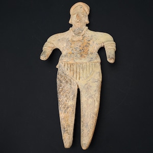 Pode incluir: Uma estatueta de cor bege, com aspecto antigo, de uma pessoa com os braços estendidos. A figura tem um chapéu, um rosto detalhado e usa uma parte de cima e calças. A estatueta é feita de um material áspero e tem uma aparência desgastada.