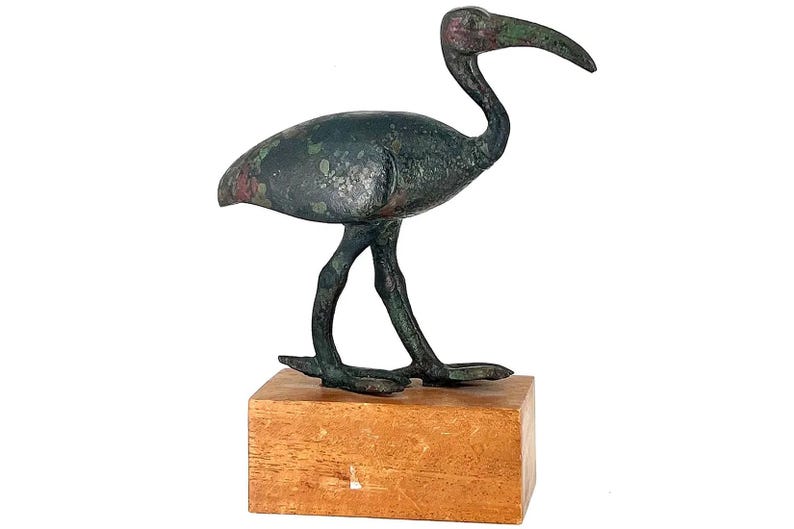 Peut inclure: Une sculpture d'oiseau en bronze avec un long bec incurv&eacute;, pos&eacute;e sur un bloc de bois. L'oiseau pr&eacute;sente une patine vert fonc&eacute; avec des reflets rouges. La sculpture est un objet d&eacute;coratif.