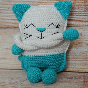 May include: A crocheted white and turquoise cat toy with a smiling face. The cat is holding its arms in front of its chest.