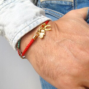 May include: A red braided bracelet with a gold charm of a hand and a horseshoe. The bracelet is worn on a person's wrist.