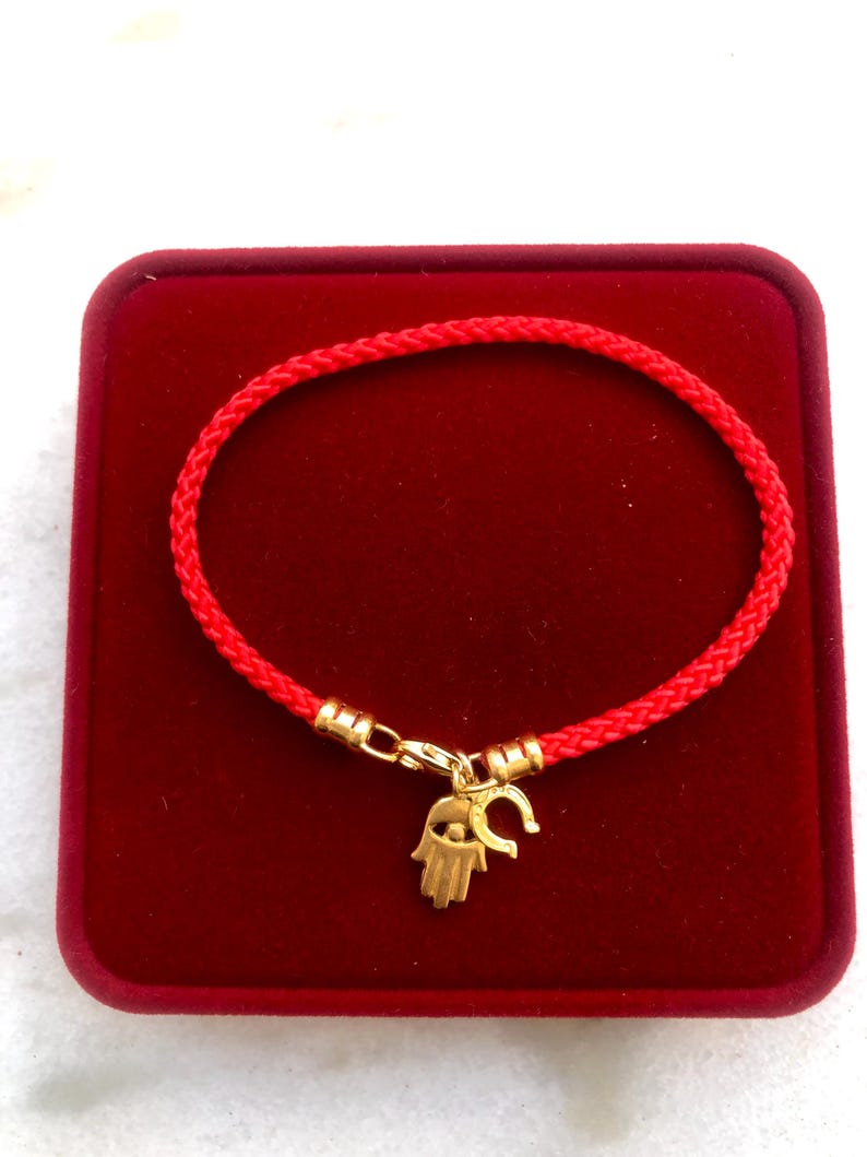 May include: Red string bracelet with a gold charm featuring a hamsa hand with an evil eye and a horseshoe.