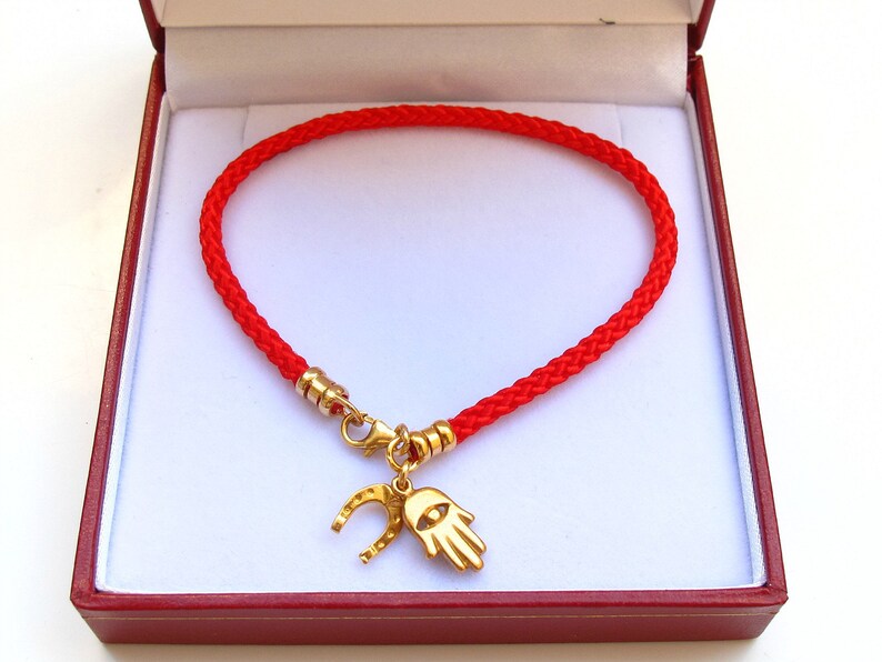 May include: Red string bracelet with a gold charm featuring a hamsa hand and horseshoe. The bracelet is in a red velvet box.