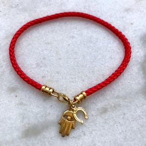 May include: Red braided cord bracelet with a gold charm featuring a hamsa hand with an evil eye and a horseshoe.
