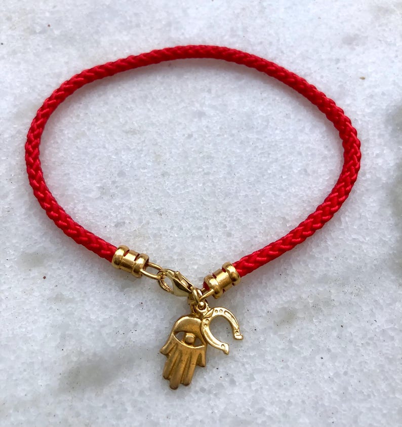 May include: Red braided cord bracelet with a gold charm featuring a hamsa hand with an evil eye and a horseshoe.