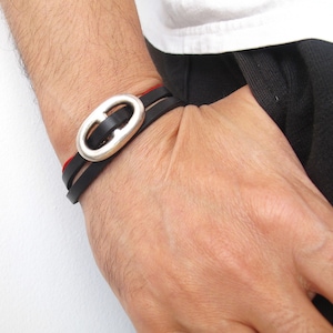 May include: A black leather bracelet with a silver oval clasp and a red leather accent.