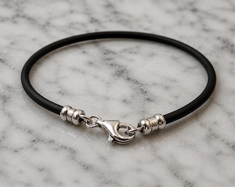 May include: A black rubber cord bracelet featuring a polished silver-tone metal lobster claw clasp and two matching silver-tone metal end caps that secure the cord on both sides of the closure mechanism.