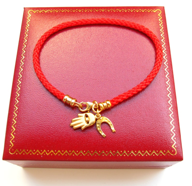 May include: A red string bracelet with a gold charm featuring a hamsa hand and a horseshoe. The bracelet is displayed in a red velvet box.