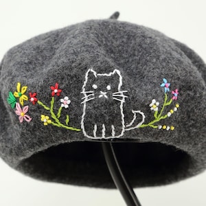 May include: A dark gray beret with an embroidered design. The design features a white cat and colorful flowers. The flowers include red, pink, yellow, and blue. The beret is made of a soft, felt-like material.