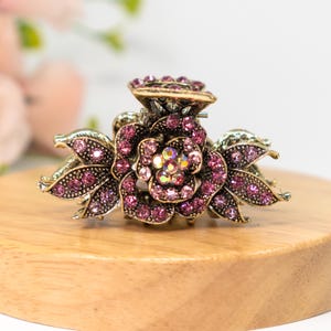May include: A gold-tone hair clip featuring a pink rhinestone flower design. The clip is adorned with small, sparkling rhinestones.