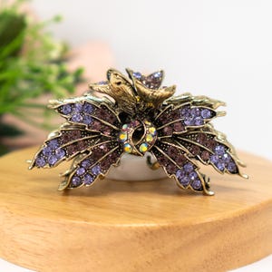 Op de afbeelding: Een gouden haarspeld met een bloemmotief. De speld is versierd met paarse en iriserende strass steentjes.