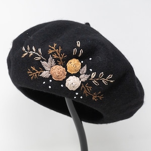 May include: A black beret featuring intricate embroidery of three circular floral motifs in shades of cream, tan, and gold, surrounded by stitched leafy branches and small white dots, creating a detailed botanical pattern on the front surface.