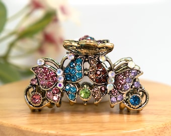 May include: A gold-tone hair clip featuring a butterfly design. The clip is adorned with colourful rhinestones in shades of pink, blue, green, and brown.