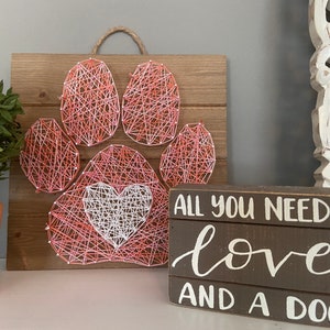 May include: A pink and white string art paw print with a heart in the center. The paw print is on a wooden board. A wooden sign below reads "All you need is love and a dog".