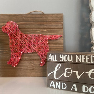 May include: A wooden sign with the text "All you need is love and a dog" in white lettering. A red string art piece of a dog is displayed on the left side of the image.