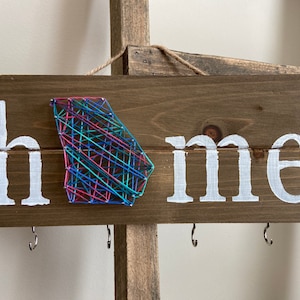 May include: A wooden sign with the word "home" painted in white. The letter "o" is replaced with a string art design of the state of Georgia in pink, blue, and green thread.