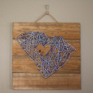 May include: A wooden sign with a blue and white string art design in the shape of the state of South Carolina with a heart in the middle.