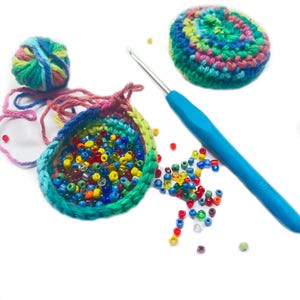 May include: A colorful crochet project in progress, featuring a partially completed round with a rainbow yarn edge, filled with assorted beads. A blue-handled crochet hook and a ball of yarn are also present, along with a finished crochet circle.