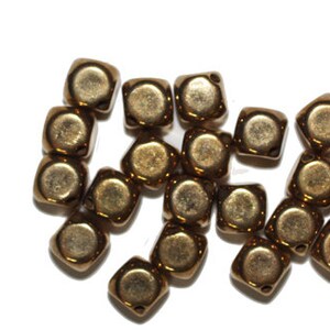 Puede incluir: Un grupo de cuentas cuadradas de color dorado. Las cuentas son de vidrio y tienen un acabado brillante.