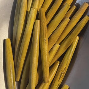 May include: A collection of numerous, tapered, cylindrical wooden dowels. The dowels are a muted yellow color with visible wood grain. The ends are flat and the overall shape is smooth. The dowels are arranged in a pile.