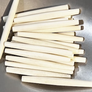 May include: A pile of light beige wooden dowels, each with a rounded body and tapered ends. The dowels are arranged in a slightly overlapping pattern, suggesting a craft or woodworking project. The natural wood color provides a neutral, earthy tone.