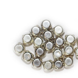 May include: A pile of silver-colored metal beads, each with a square shape and a rounded top. The beads are arranged in a loose cluster.