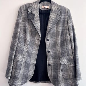 Puede incluir: Un blazer a cuadros grises con interior negro y tres botones negros. El blazer tiene dos bolsillos delanteros y está colgado de una percha negra. Una etiqueta está adherida a la solapa.