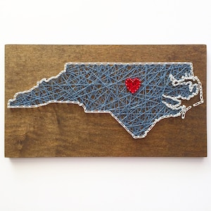 May include: A string art piece depicting the outline of North Carolina with a red heart in the center. The art is on a wooden background.