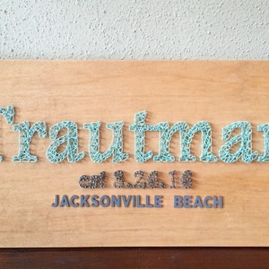 Op de afbeelding: Een houten bord met de naam "Trautman" gespeld in lichtblauwe string art. De tekst "est. 8.26.16" en "JACKSONVILLE BEACH" staan onder de naam in grijze string art.