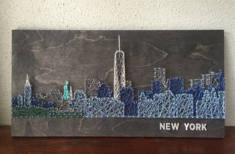 K&ouml;nnte beinhalten: Eine String-Art-Darstellung der Skyline von New York City, mit dem ikonischen Freedom Tower und anderen Wolkenkratzern. Das Kunstwerk ist mit blauem, wei&szlig;em und gr&uuml;nem Garn auf einem dunklen Holzgrund geschaffen. Die Worte "NEW YORK" sind in wei&szlig;en Buchstaben am unteren Rand des Kunstwerks geschrieben.