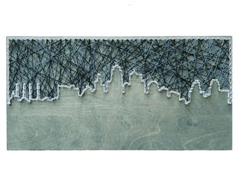 May include: A string art piece depicting a city skyline. The skyline is outlined in white string and filled in with black string. The background is a light grey wood.