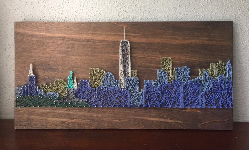 K&ouml;nnte beinhalten: Ein String-Art-Bild, das die Skyline von New York City darstellt, mit dem ikonischen Freedom Tower und anderen Wolkenkratzern. Das Kunstwerk wird mit bunten F&auml;den geschaffen, die zwischen N&auml;geln gespannt sind, die in eine Holzplatte geh&auml;mmert wurden.