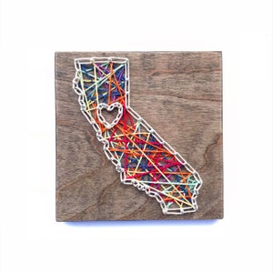 May include: String art of the state of California with a small heart in the center. The art is made with colorful string wrapped around nails hammered into a wooden board.