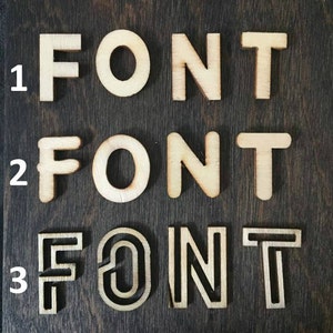 K&ouml;nnte beinhalten: Drei verschiedene Stile des Wortes "FONT" aus Holz ausgeschnitten. Der erste Stil ist eine einfache, klobige Schriftart. Der zweite Stil ist eine leicht abgerundete Schriftart. Der dritte Stil ist eine dekorativere Schriftart mit einer doppelten Umrandung.