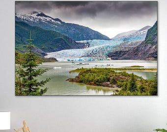 May include: A rectangular wall art print featuring a landscape of a blue glacier flowing between dark mountains into a calm lake, with green pine trees in the foreground under a cloudy sky, mounted above a light brown wooden sideboard cabinet.