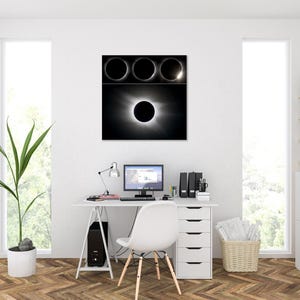 May include: A black and white framed print of a solar eclipse hangs above a white desk. The print shows the stages of an eclipse. A computer, lamp, and office supplies sit on the desk. A white chair and a potted plant are also in the room.