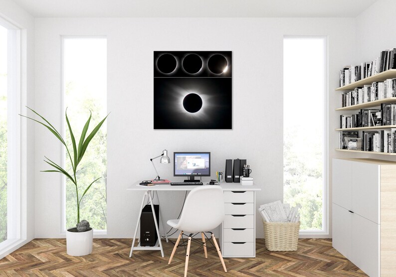 May include: A black and white framed print of a solar eclipse hangs above a white desk. The print shows the stages of an eclipse. A computer, lamp, and office supplies sit on the desk. A white chair and a potted plant are also in the room.