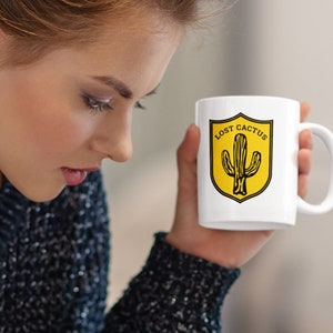 May include: White ceramic mug with a yellow and black graphic of a cactus and the text "Lost Cactus".