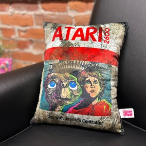May include: A decorative pillow with a vintage Atari 2600 game cartridge design. The pillow features a distressed, faded look with a graphic of a creature from the movie E.T. and a young boy. The text on the pillow reads "ATARI 2600 Use with Joystick Controller."
